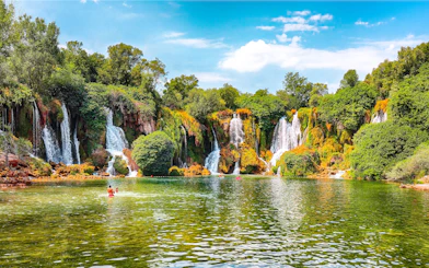 Kravice Waterfall with lush greenery and people swimming in the foreground.