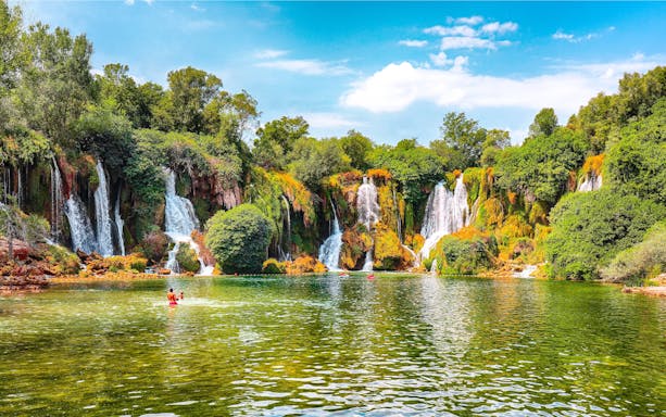 Kravice Waterfall with lush greenery and people swimming in the foreground.