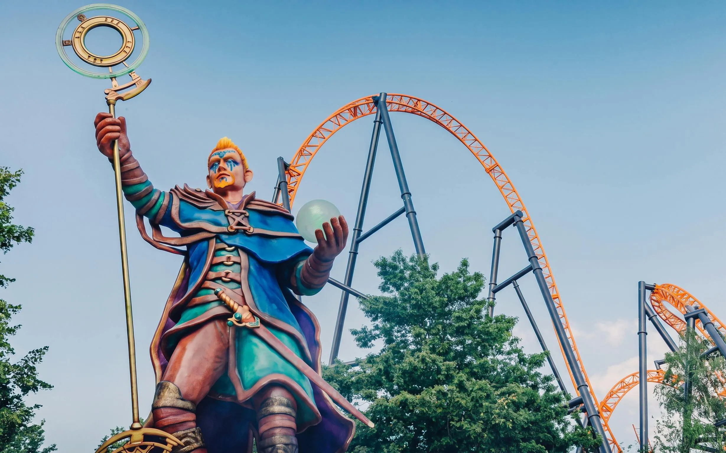 Statue of a wizard with a staff and orb at Bobbejaanland's Land of Legends, roller coaster in background.