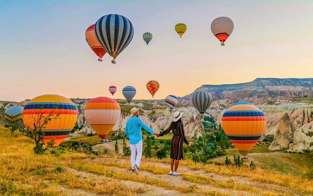 Couple holding hands watching hot air balloons over Cappadocia landscape.