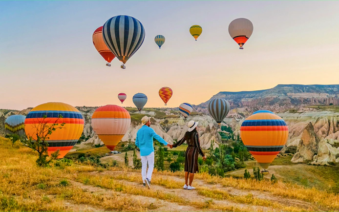 Couple holding hands watching hot air balloons over Cappadocia landscape.