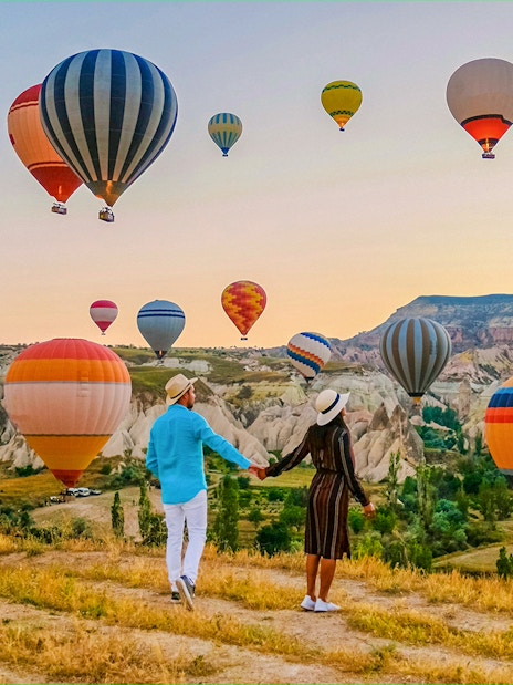 Couple holding hands watching hot air balloons over Cappadocia landscape.