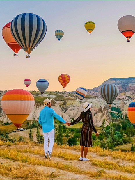 Couple holding hands watching hot air balloons over Cappadocia landscape.
