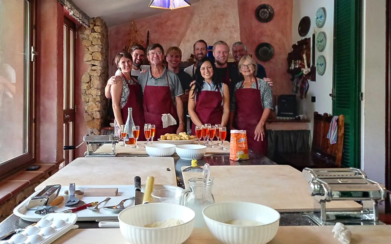 Group in aprons at pasta cooking class in Alghero kitchen with tools and ingredients.