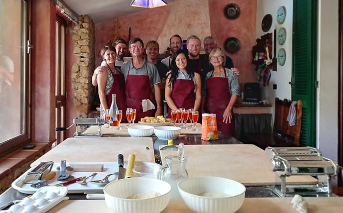 Group in aprons at pasta cooking class in Alghero kitchen with tools and ingredients.