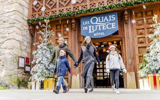 Family enjoying Christmas at Parc Asterix, walking by decorated trees and Les Quais de Lutece Hotel.
