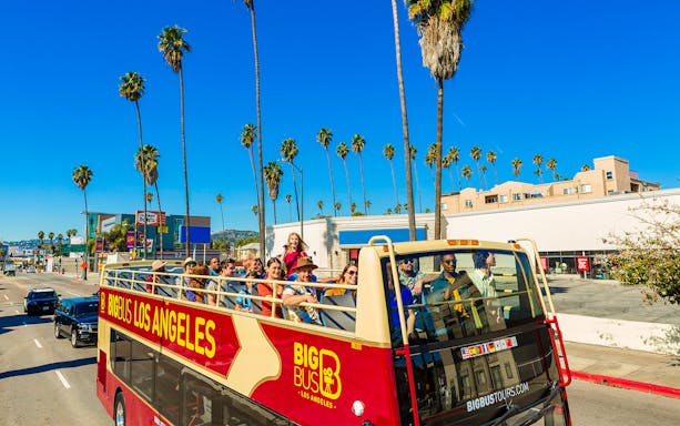 Open-top tour bus with passengers on a sunny street in Los Angeles, lined with palm trees.