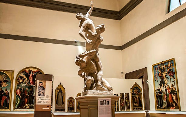 Sculpture of three figures in the Accademia Gallery, Florence, surrounded by Renaissance paintings.