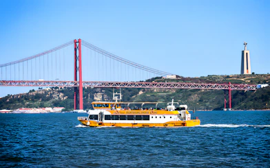 Yellow boat on Lisbon's Tagus River with 25 de Abril Bridge and Cristo Rei statue in the background.