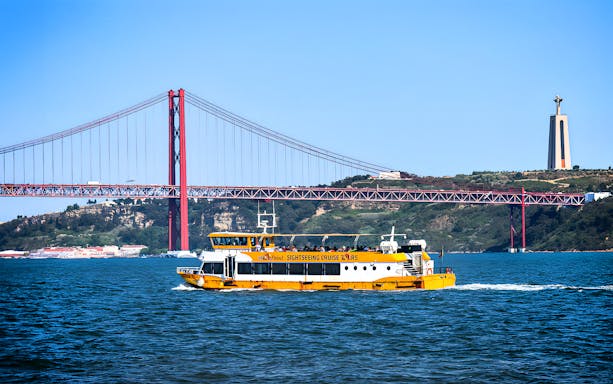 Yellow boat on Lisbon's Tagus River with 25 de Abril Bridge and Cristo Rei statue in the background.