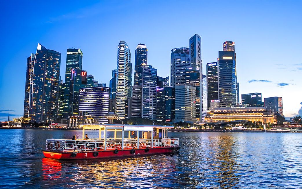 Singapore River Cruise boat on water with city skyline at dusk.