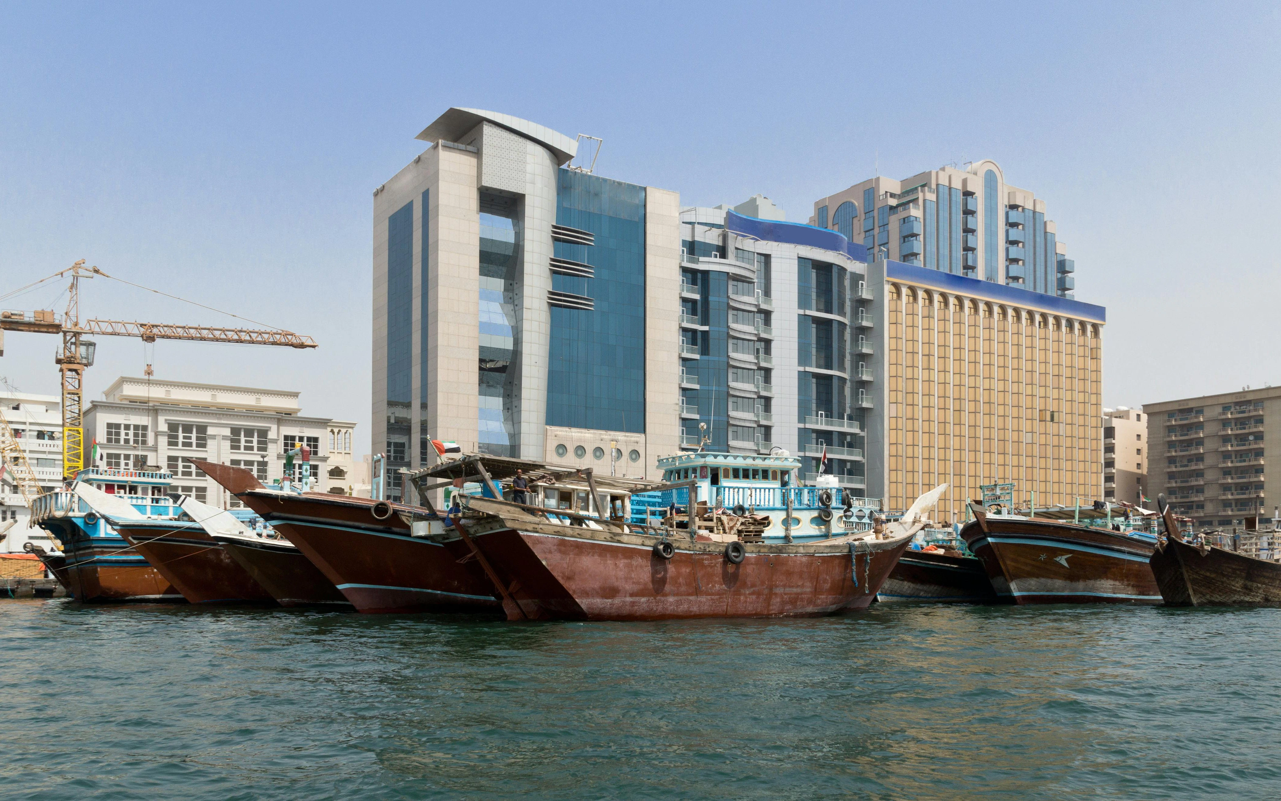 Traditional wooden dhows docked along Dubai Creek with modern buildings in the background.