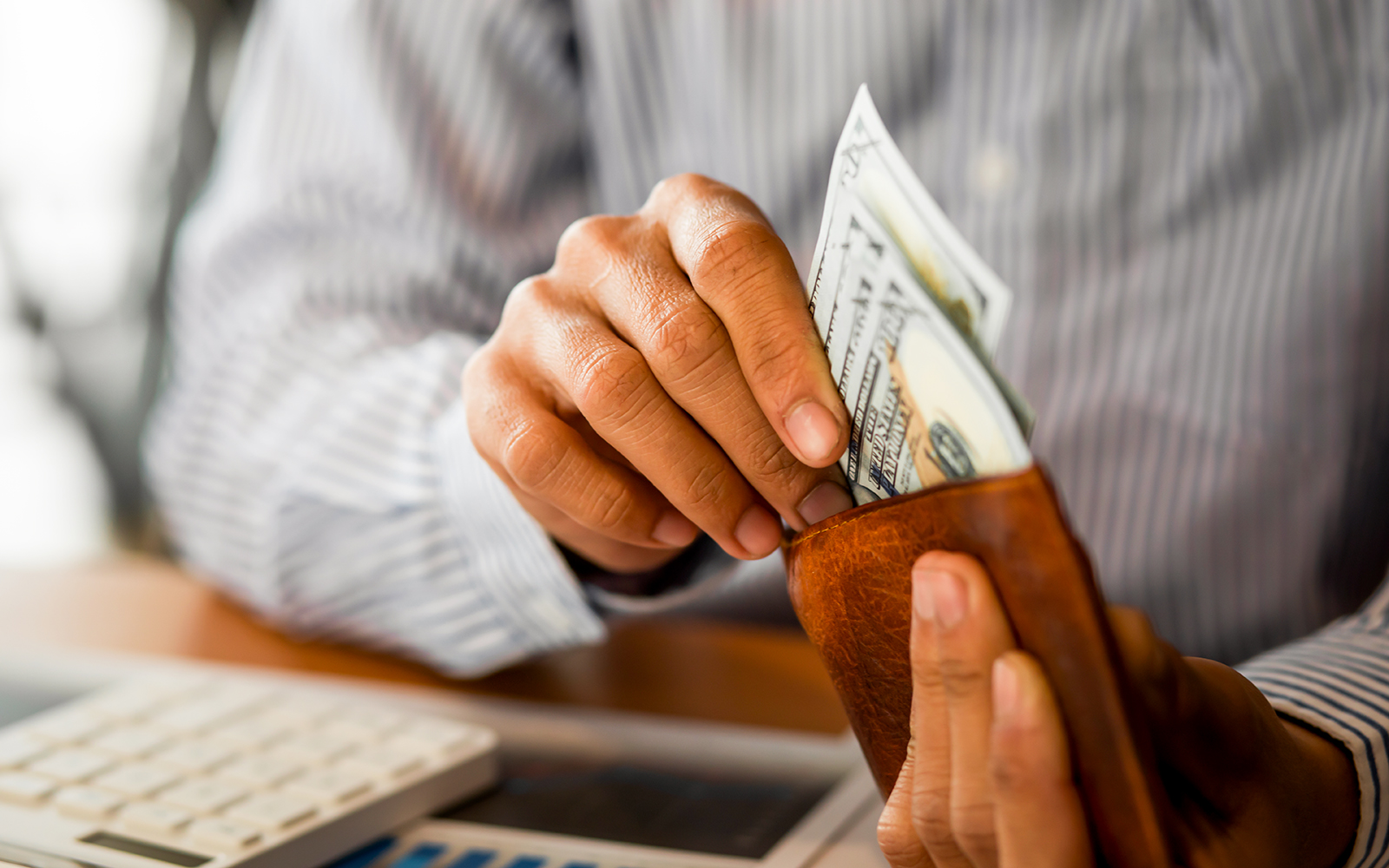 Hands placing cash into a wallet near a calculator.