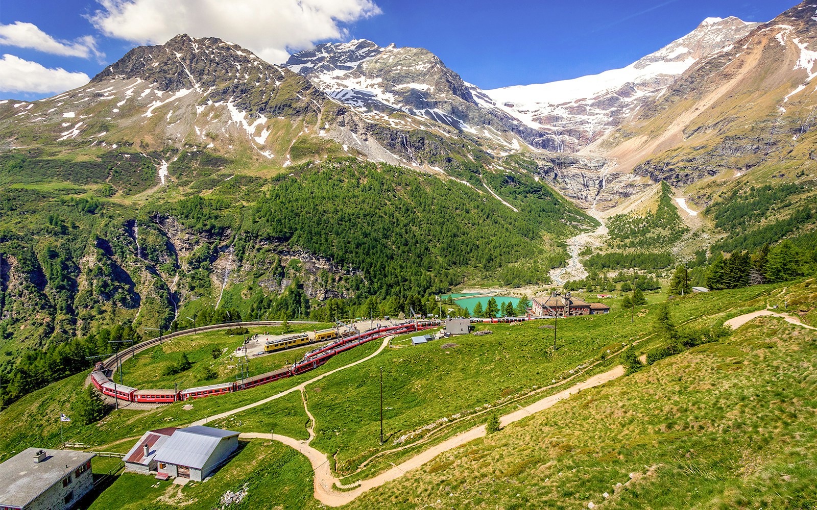 Bernina Express train at Alp Grum Station with snow-capped mountains in Switzerland.