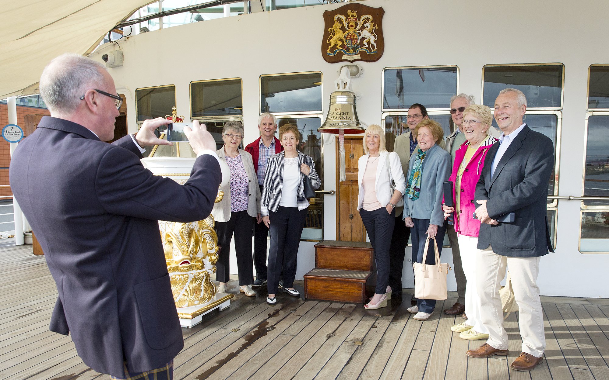 Tourists posing on the deck of a ship with a bell and crest in the background.