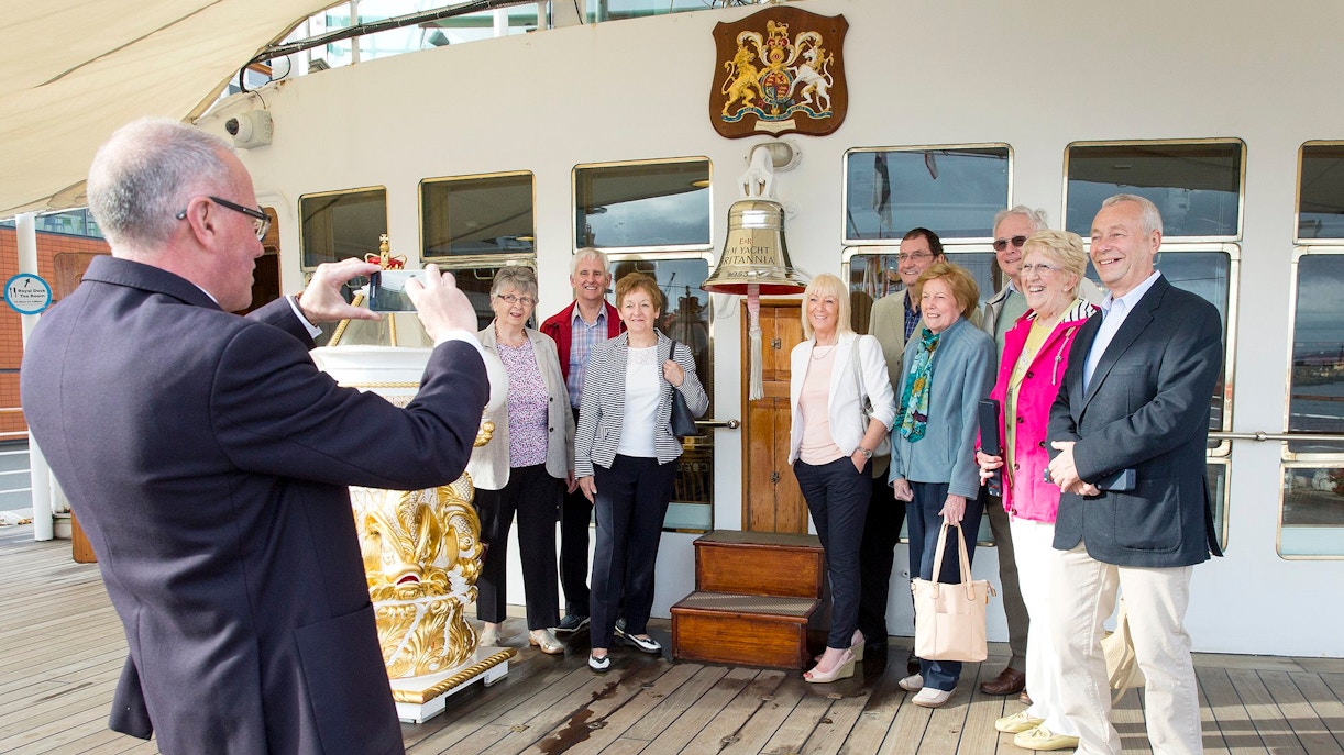 Tourists posing on the deck of a ship with a bell and crest in the background.