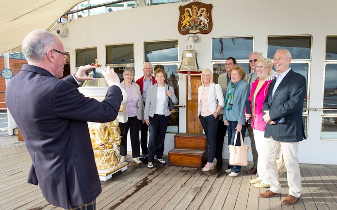 Tourists posing on the deck of a ship with a bell and crest in the background.
