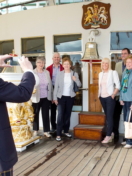 Tourists posing on the deck of a ship with a bell and crest in the background.