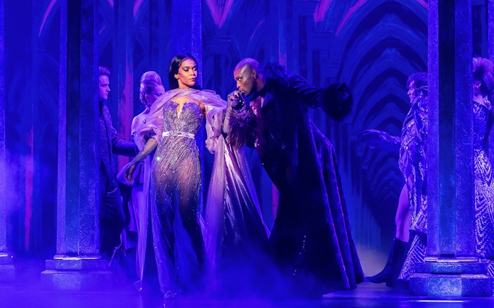 Performers in a theatrical scene from "Death Becomes Her" with dramatic lighting and costumes.