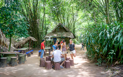 Visitors gathering at Cu Chi Tunnels, Vietnam, near a thatched hut surrounded by lush greenery.