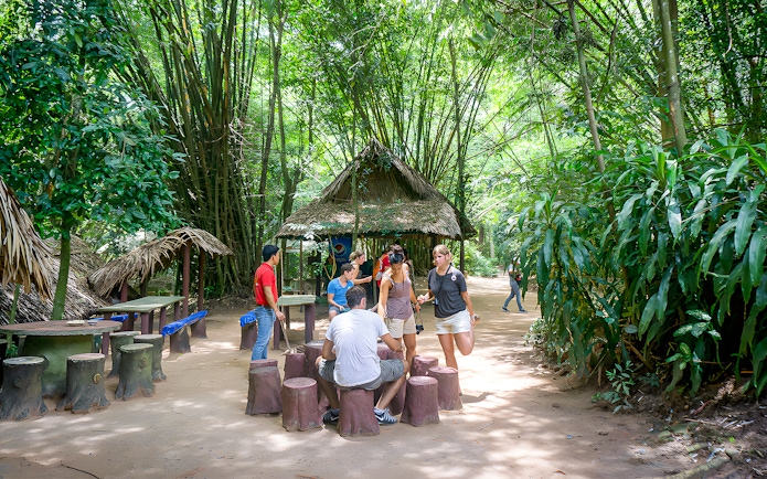 Visitors gathering at Cu Chi Tunnels, Vietnam, near a thatched hut surrounded by lush greenery.