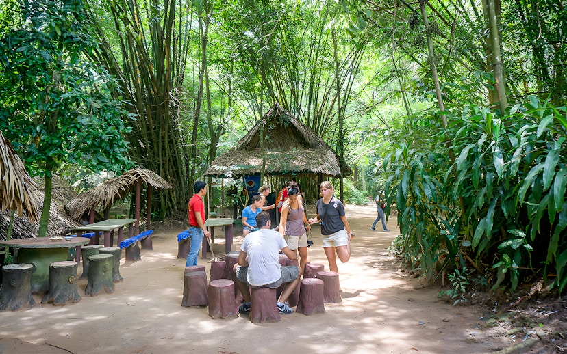 Visitors gathering at Cu Chi Tunnels, Vietnam, near a thatched hut surrounded by lush greenery.