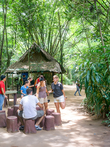 Visitors gathering at Cu Chi Tunnels, Vietnam, near a thatched hut surrounded by lush greenery.