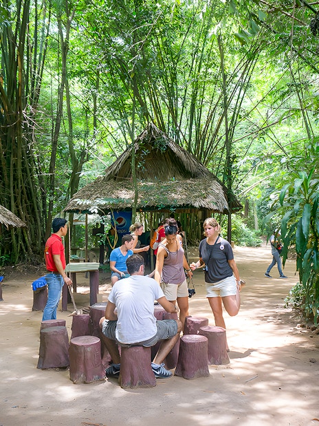 Visitors gathering at Cu Chi Tunnels, Vietnam, near a thatched hut surrounded by lush greenery.
