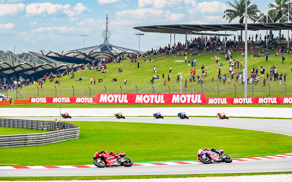 Motorcycles racing at Petronas Grand Prix of Malaysia 2023 with spectators on grassy hill.