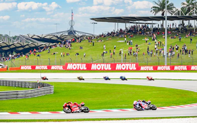 Motorcycles racing at Petronas Grand Prix of Malaysia 2023 with spectators on grassy hill.