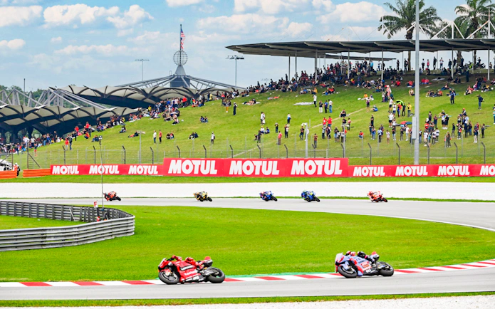 Motorcycles racing at Petronas Grand Prix of Malaysia 2023 with spectators on grassy hill.