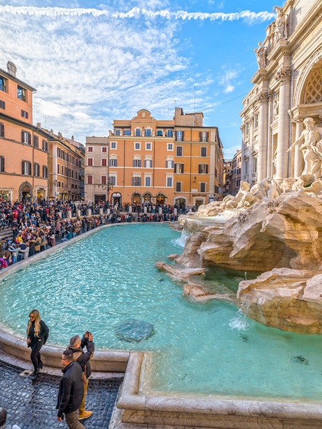 Crowd gathered around Trevi Fountain in Rome, Italy, with historic architecture in the background.