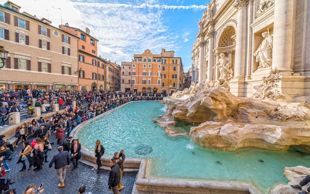 Crowd gathered around Trevi Fountain in Rome, Italy, with historic architecture in the background.