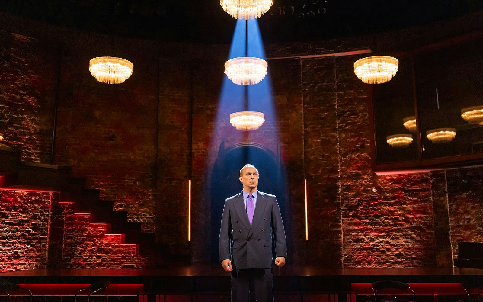 Man standing under spotlight in theater with brick walls and chandeliers, related to Patriots.