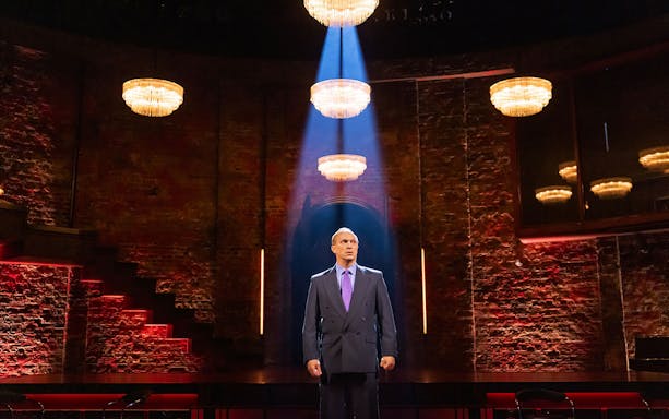 Man standing under spotlight in theater with brick walls and chandeliers, related to Patriots.
