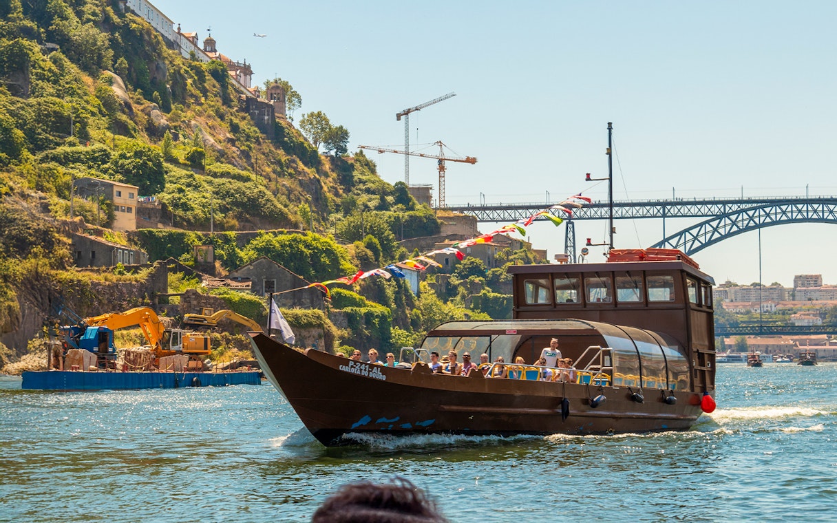 Tourists on a Douro River cruise boat with scenic hillside and bridge in Porto, Portugal.