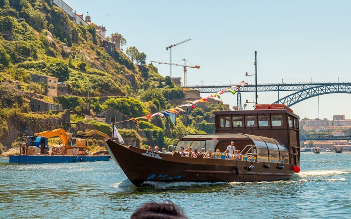 Tourists on a Douro River cruise boat with scenic hillside and bridge in Porto, Portugal.