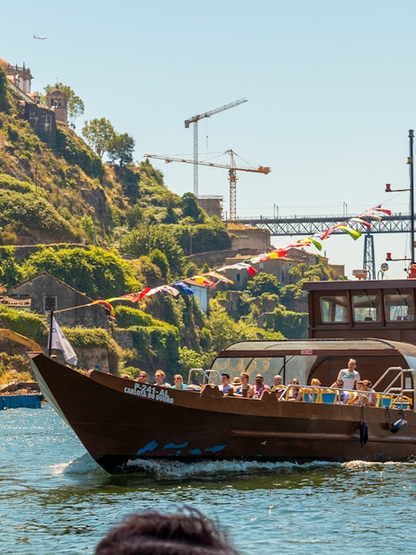 Tourists on a Douro River cruise boat with scenic hillside and bridge in Porto, Portugal.