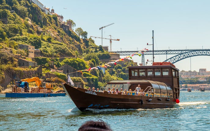 Tourists on a Douro River cruise boat with scenic hillside and bridge in Porto, Portugal.