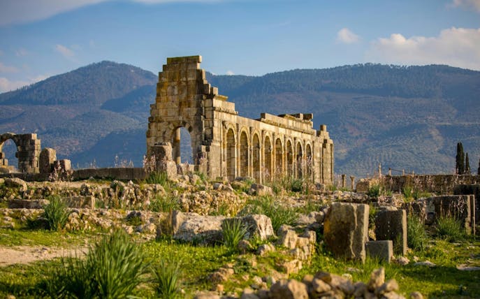 Volubilis Roman ruins with arches and columns in Fes, Morocco, against a mountain backdrop.