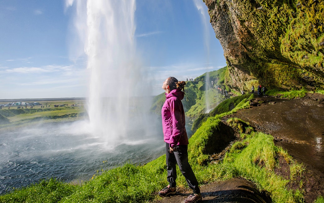Woman standing near Seljalandsfoss waterfall, Iceland, with lush green surroundings.