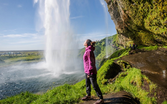 Woman standing near Seljalandsfoss waterfall, Iceland, with lush green surroundings.