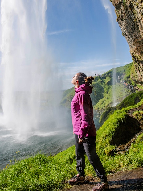 Woman standing near Seljalandsfoss waterfall, Iceland, with lush green surroundings.