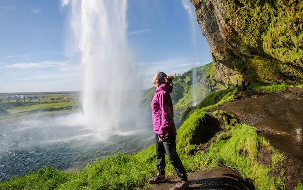 Woman standing near Seljalandsfoss waterfall, Iceland, with lush green surroundings.