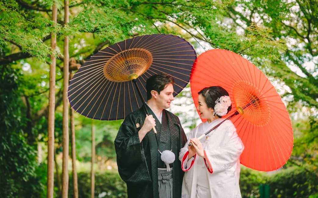 Couple in traditional attire with umbrellas during a tea ceremony experience in a Japanese garden.