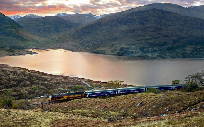 Train traveling along a scenic lake in the Scottish Highlands at sunset, part of the BritRail network.