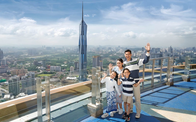 Family enjoying view from KL Tower Sky Deck, Kuala Lumpur skyline in background.