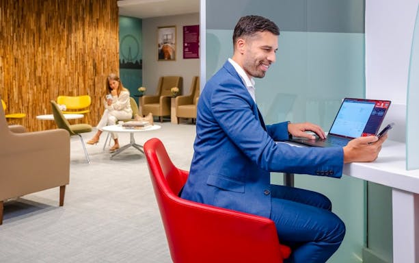 Man working on laptop in Plaza Premium Lounge, London, with seating area in background.