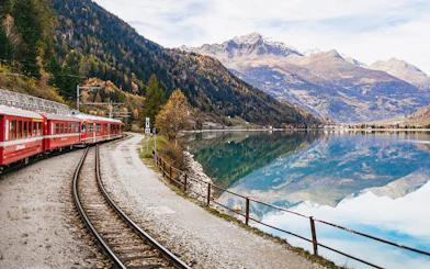 Red Bernina train traveling along a lake with mountain reflections near St. Moritz.