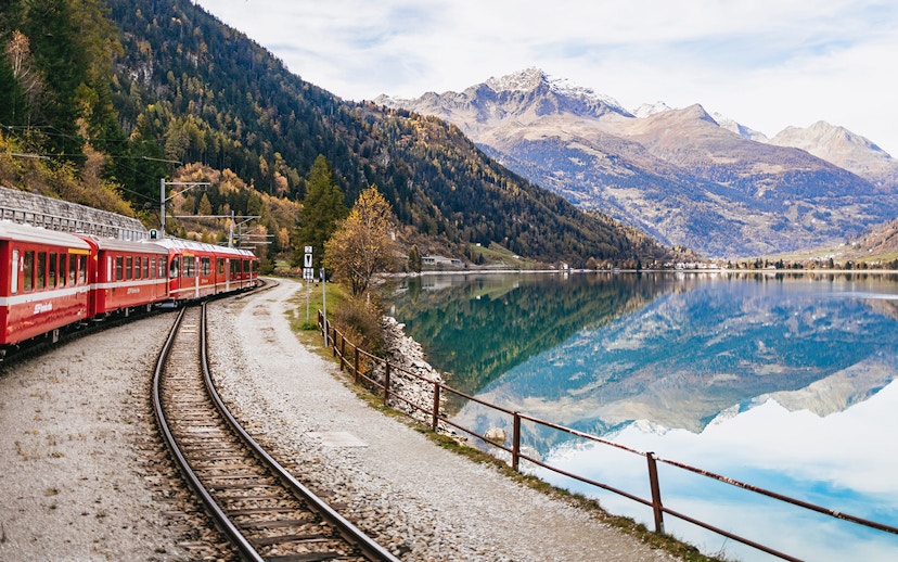 Red Bernina train traveling along a lake with mountain reflections near St. Moritz.